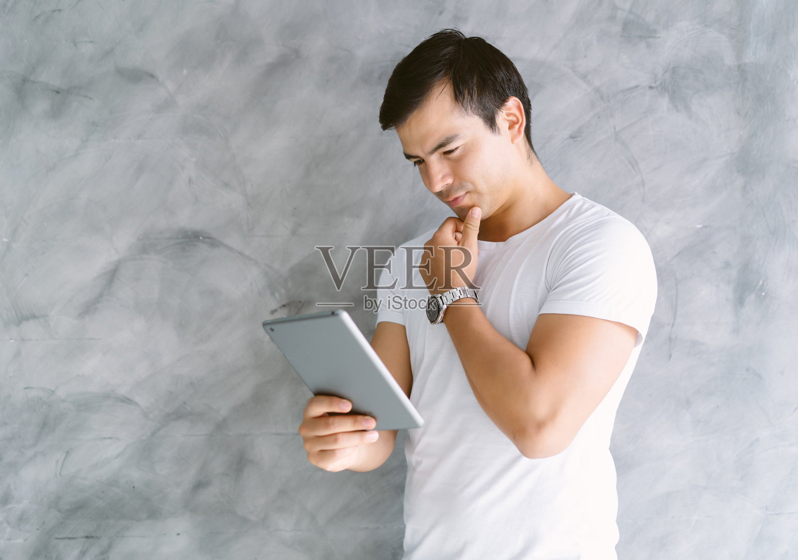 身穿白色T恤的英俊男子，他一边思考一边触摸着下巴，手里操作着平板电脑。灰色的背景和沉着的姿态传达出专注、科技应用、解决问题和现代数字工作的意境。照片摄影图片