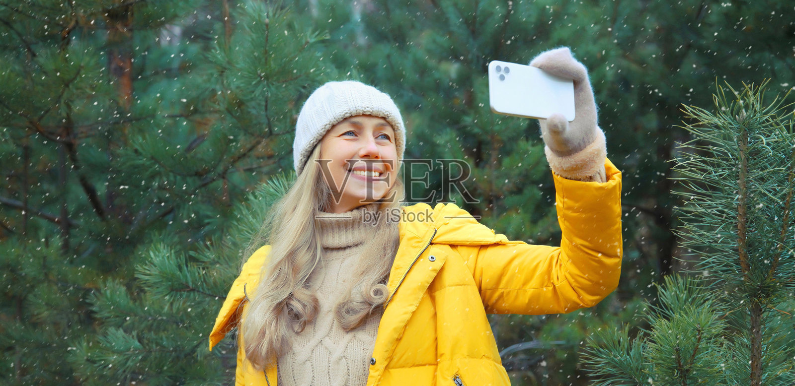 快乐微笑的女性在冬季公园里，以圣诞树为背景，用智能手机自拍照片摄影图片