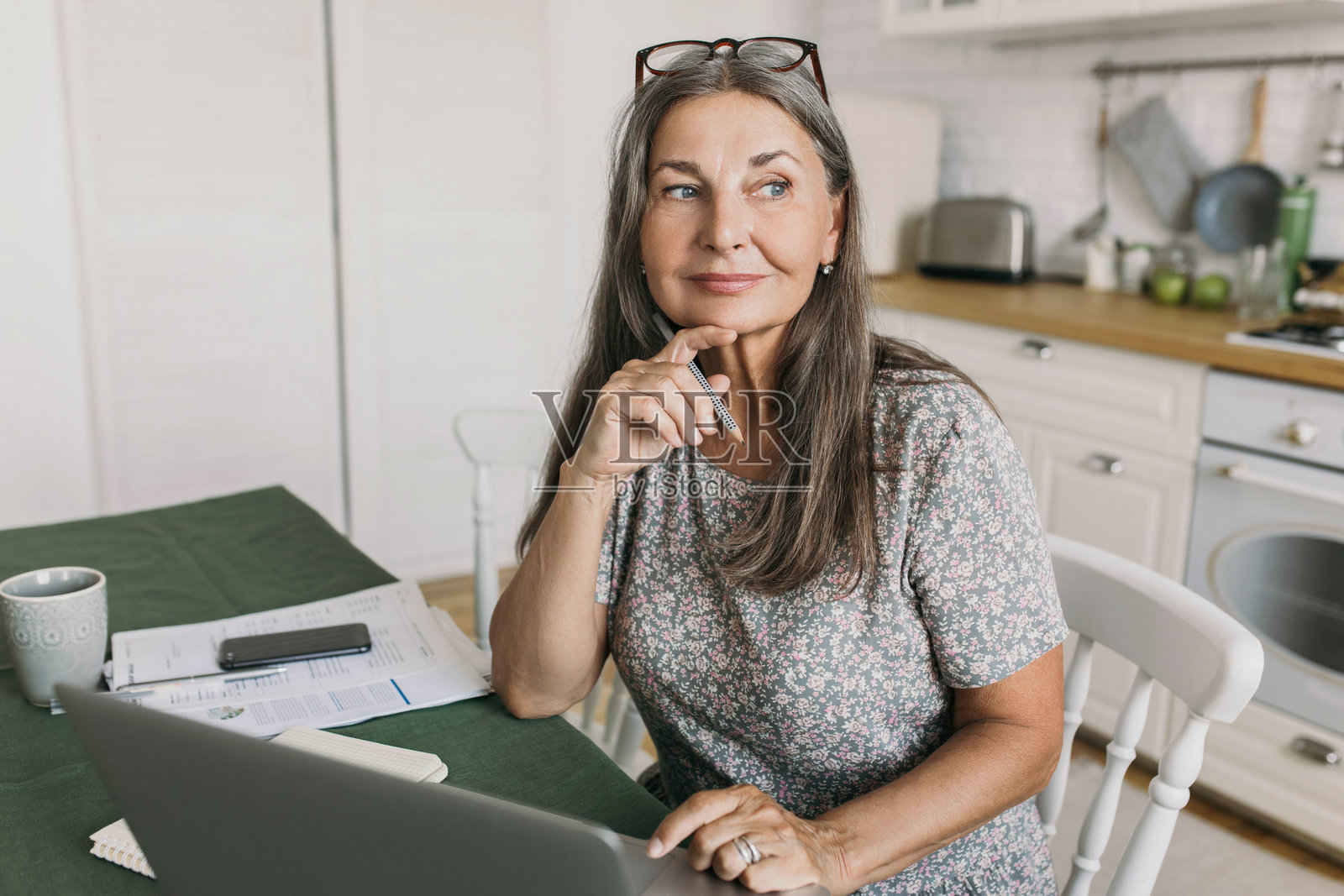 迷人的现代女作家手持钢笔，目光瞥向一旁，在厨房的笔记本电脑、手机和纸张前，为新小说构思人物情节线。创意，居家办公。照片摄影图片