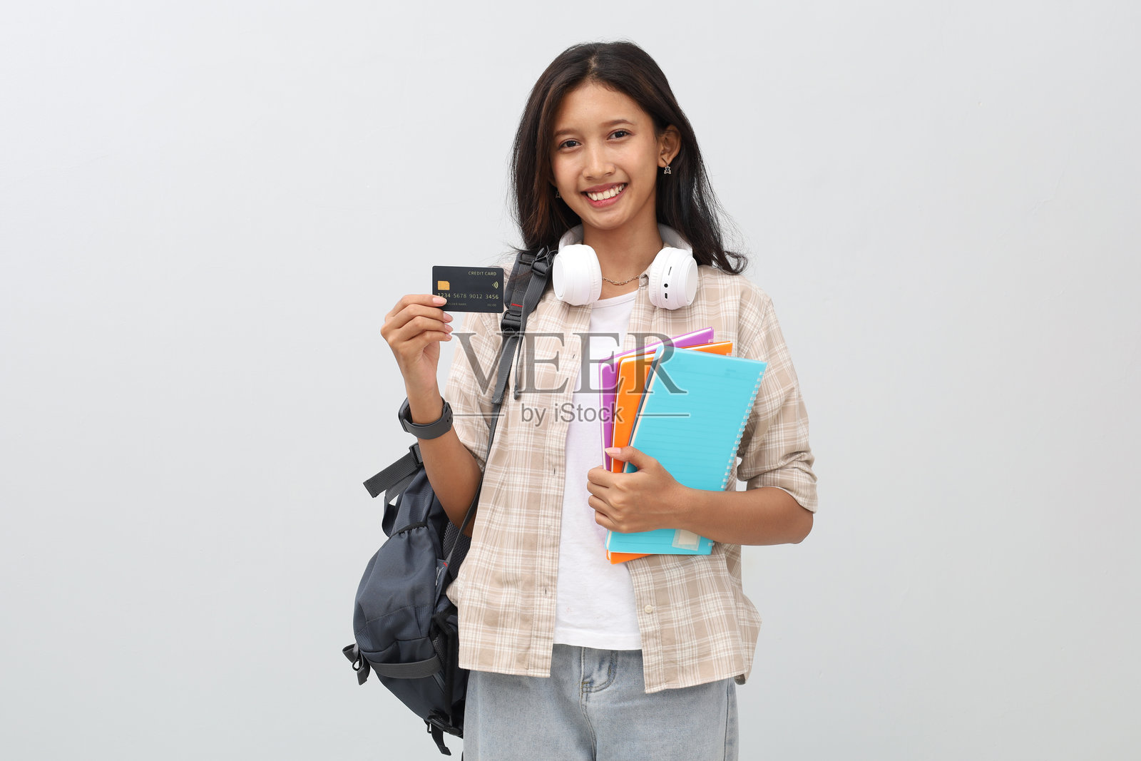 年轻的亚洲女学生穿着休闲服，背着背包，脖子上挂着耳机，手里拿着书，微笑着看向镜头，展示着信用卡，孤立在白色背景上。照片摄影图片