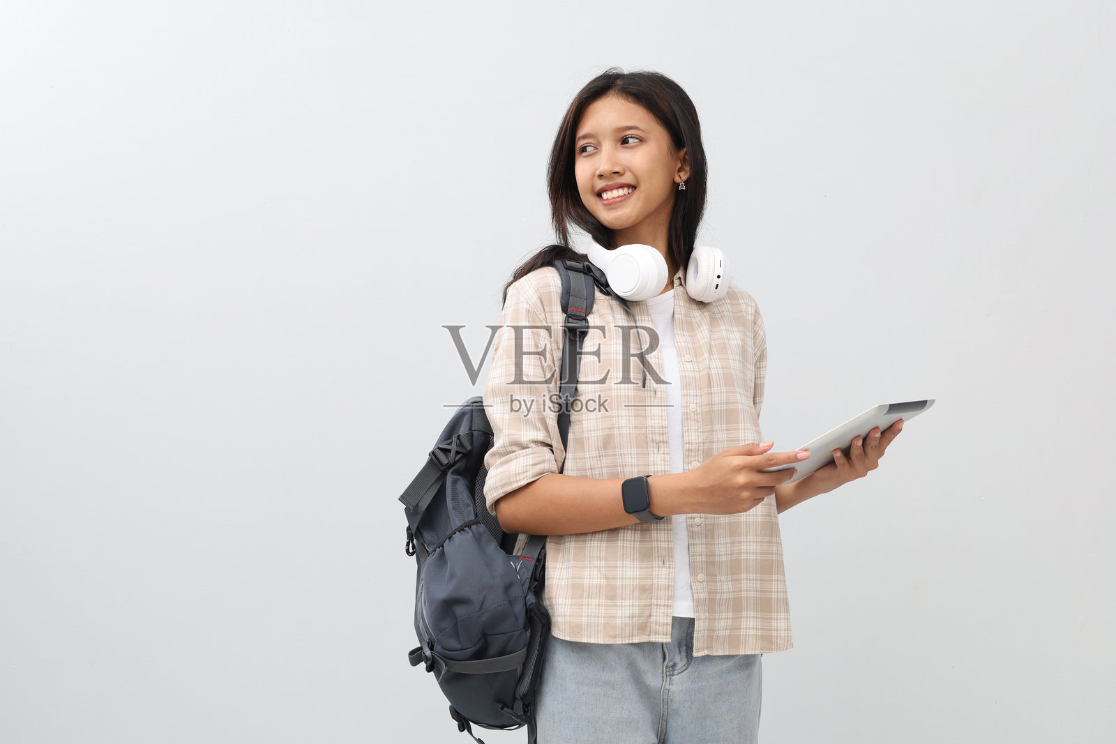 年轻的亚洲女学生，穿着休闲服，背着背包，脖子上挂着耳机，微笑着使用平板电脑，看向一旁空白处。照片摄影图片