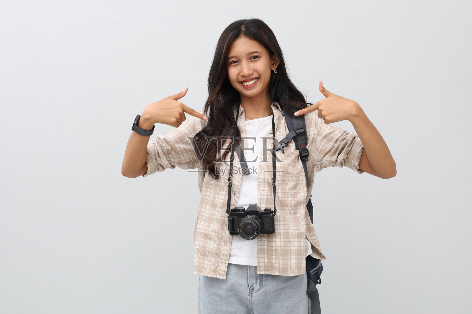 一位亚洲女性，穿着休闲服饰，背着背包，脖子上挂着相机，用食指指向自己，背景为白色，度假概念。照片摄影图片