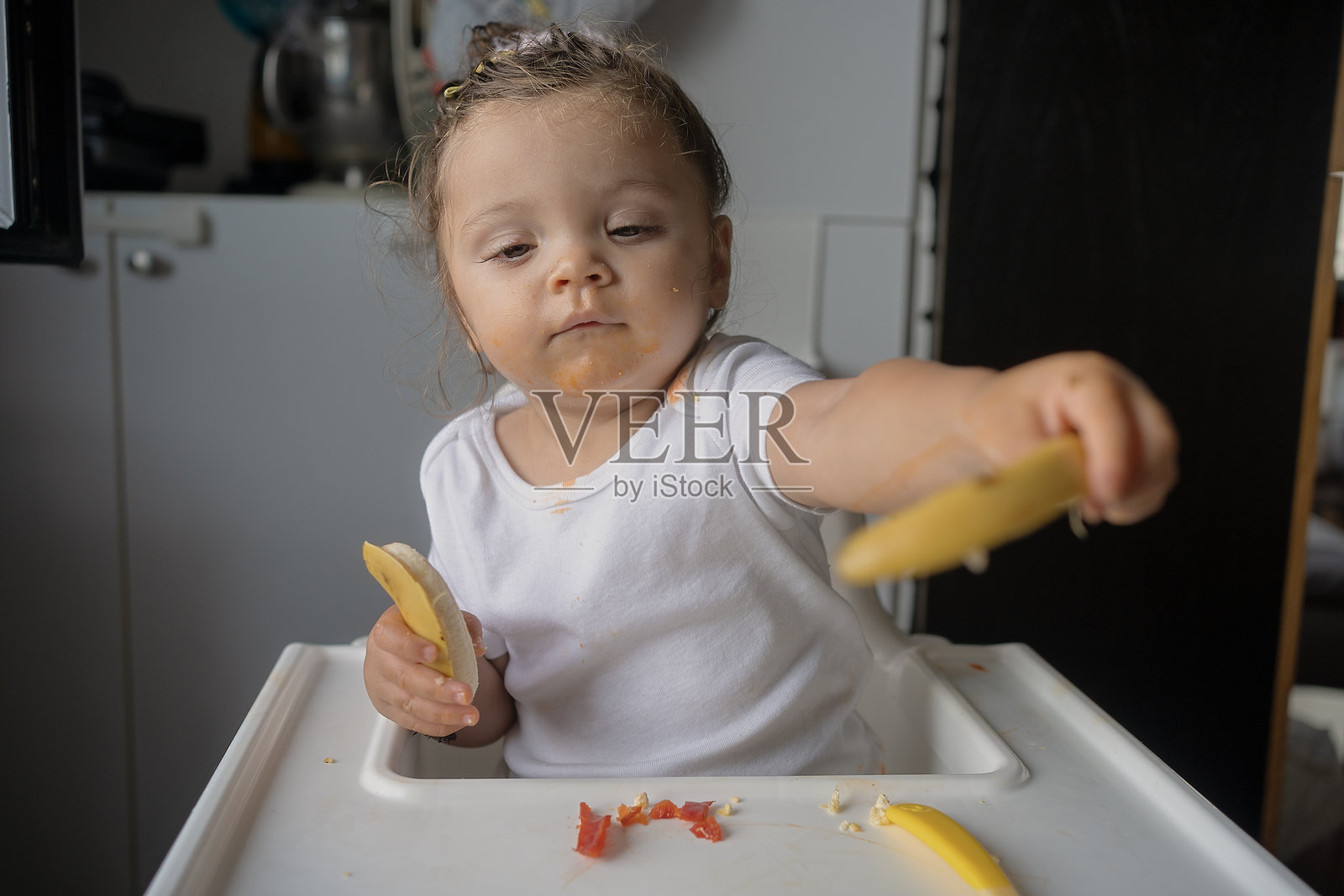 可爱的小女孩在家里坐在高脚椅上吃香蕉，弄得一团糟照片摄影图片