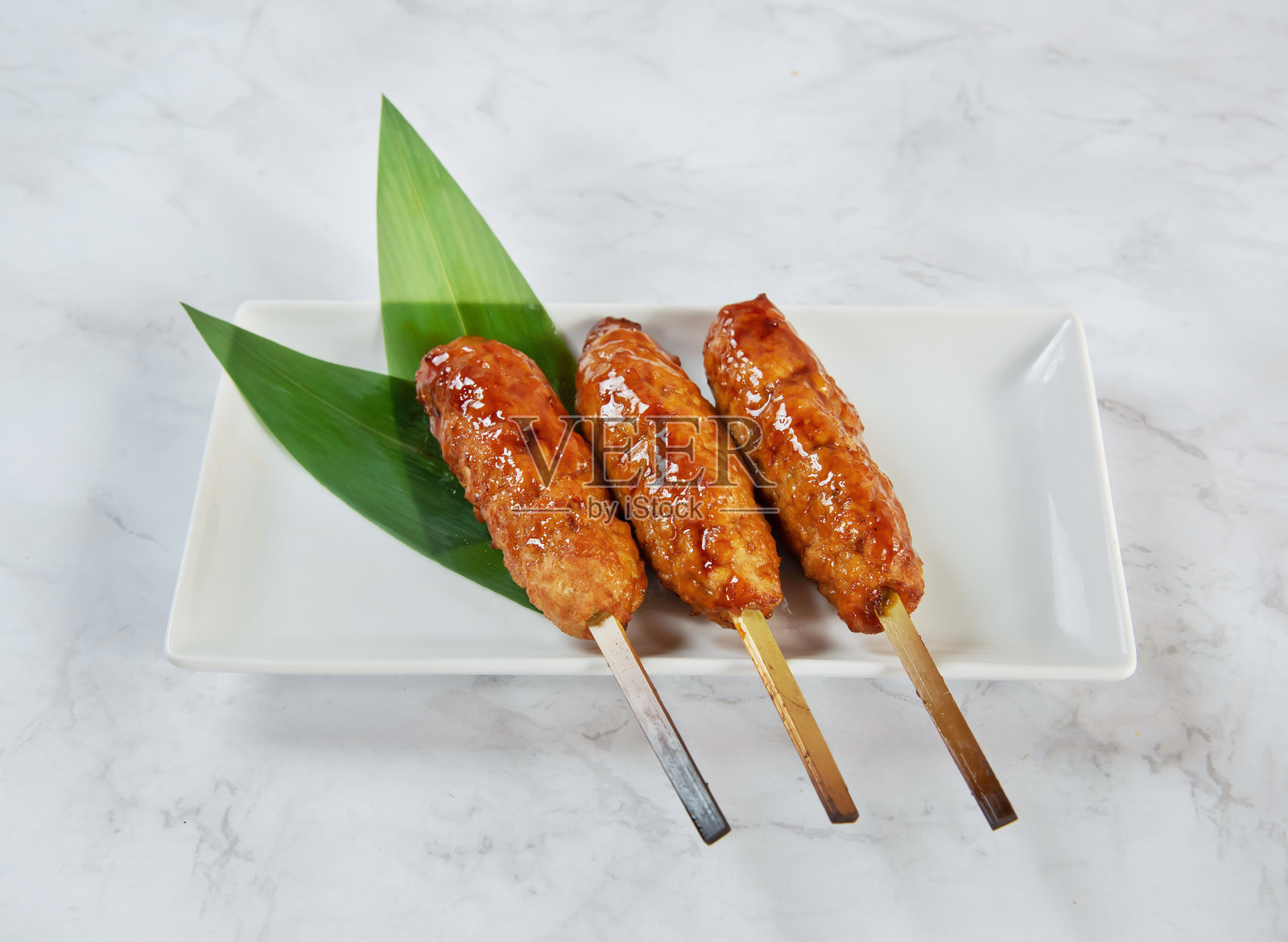 白色盘子里的釉面肉串，上面装饰着绿叶，在白色大理石背景上孤立。照片摄影图片