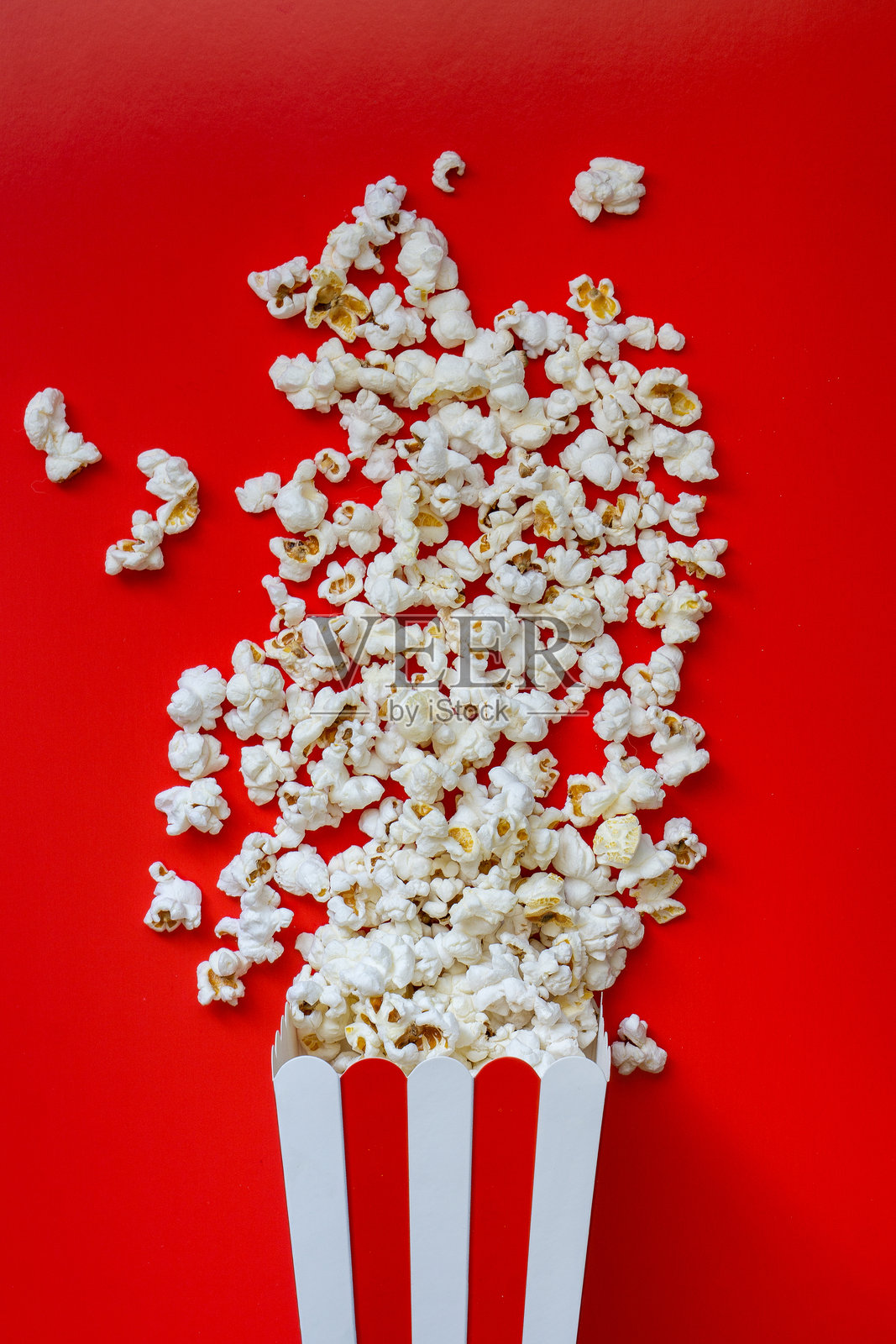 蓬松的白色爆米花从条纹纸盒中撒出，背景为鲜艳的红色。照片摄影图片