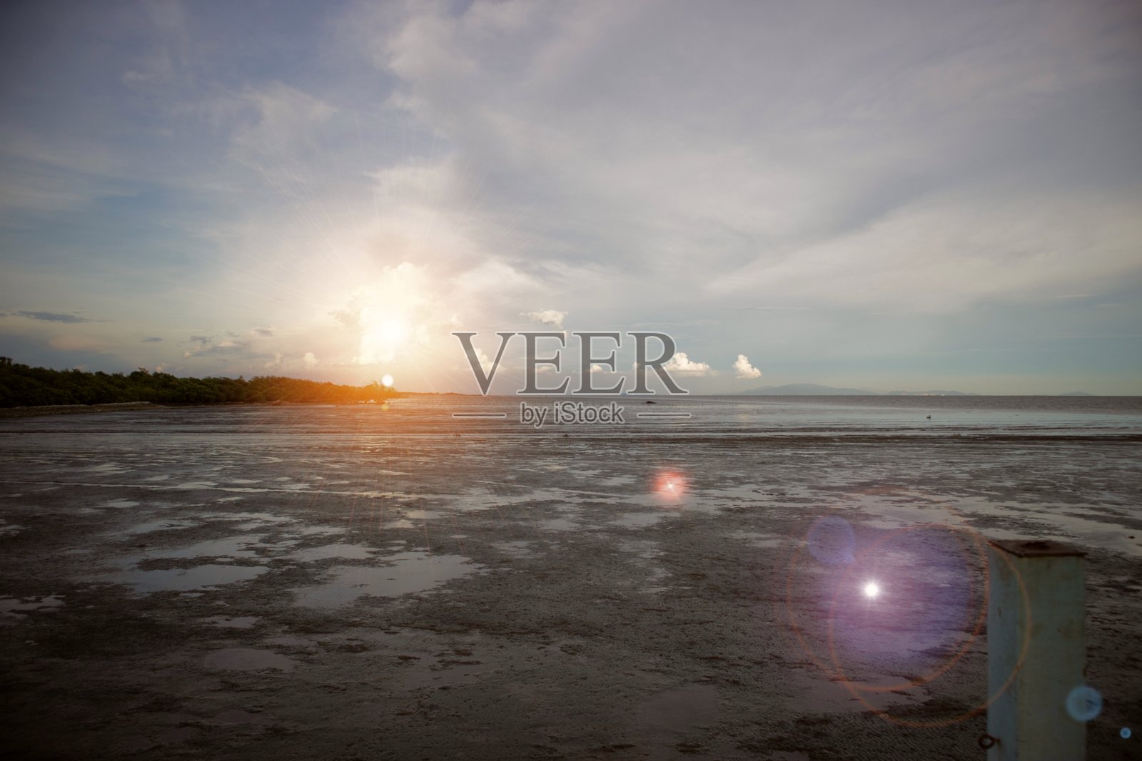 低潮时平静的海滨风光，太阳接近地平线，天空中漂浮着柔和的云彩，湿沙滩上有浅浅的水面倒影，柔和的镜头光晕营造出宁静自然的氛围。照片摄影图片