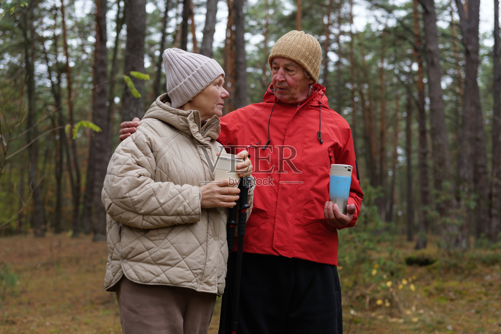 一对老年夫妇在秋天的森林里，手里拿着饮料交谈，周围是高大的树木和落叶。照片摄影图片