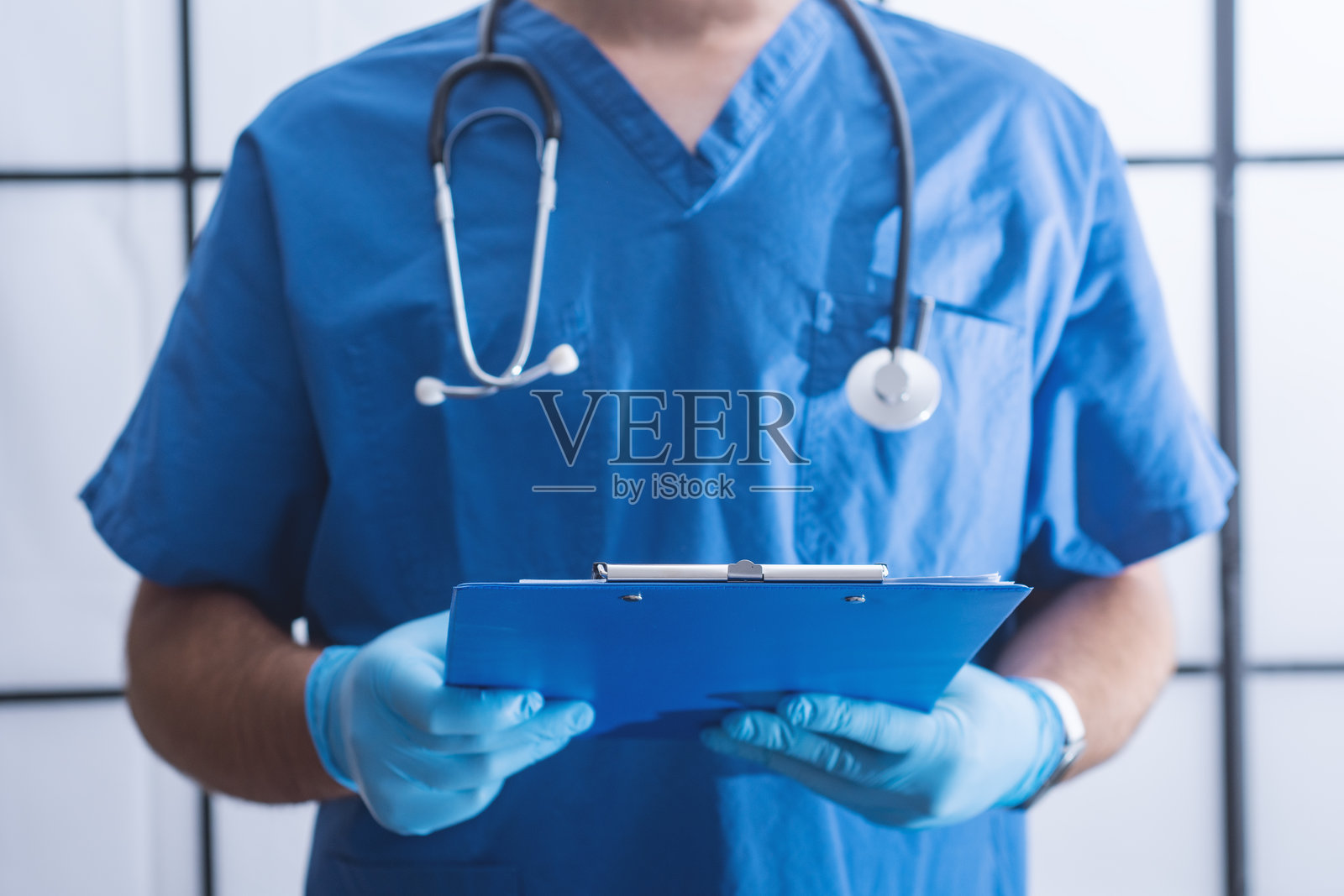 身穿蓝色手术服、戴着听诊器的男医生，在医院环境中手持蓝色病历夹，记录着病人信息。体现了医疗保健、医学咨询和值得信赖的病人护理的理念。照片摄影图片
