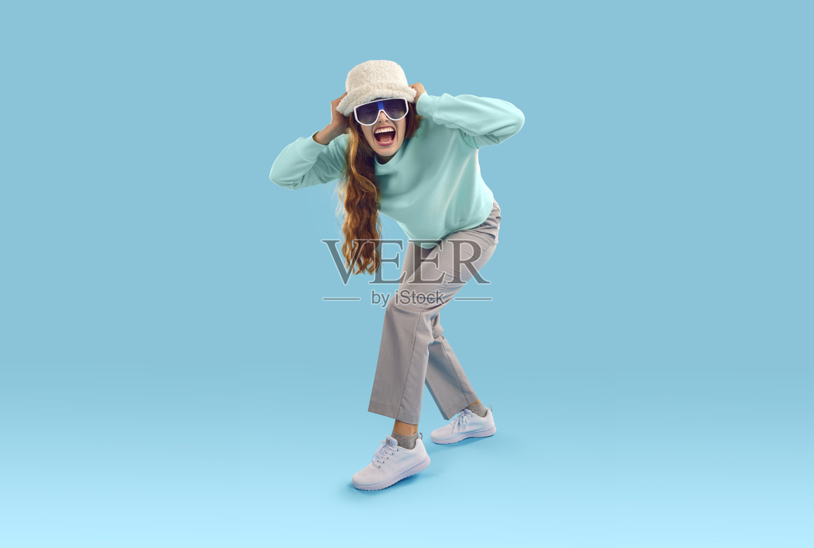 戴着帽子和太阳镜的兴奋女人在蓝色背景前尖叫照片摄影图片