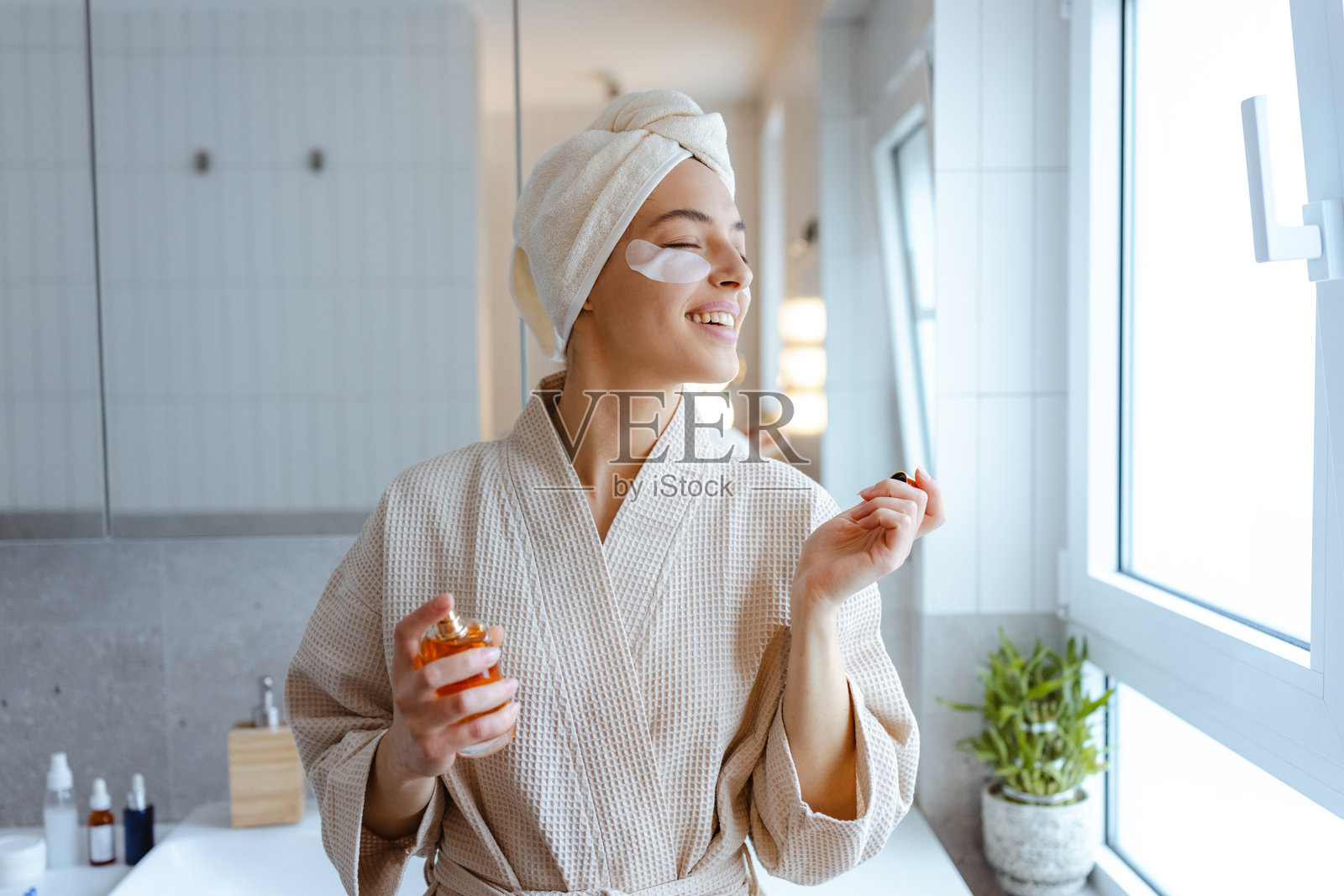 一位年轻女子在明亮的浴室里，身着浴袍，在晨间例行活动中涂抹香水照片摄影图片