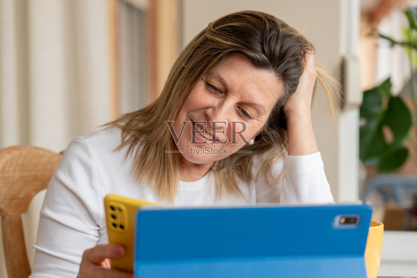 一位成熟的成年女性在家中，一边使用平板电脑和智能手机进行在线浏览和娱乐，一边露出微笑。照片摄影图片