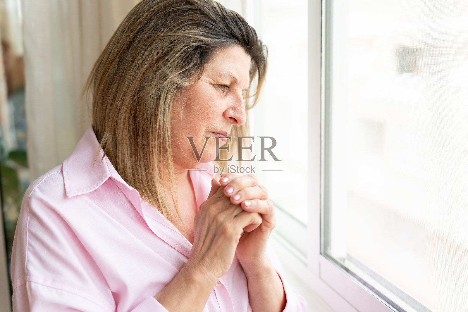 一位年长的女性独自站在窗边，双手紧握，若有所思，表达着孤独、抑郁和精神压力照片摄影图片