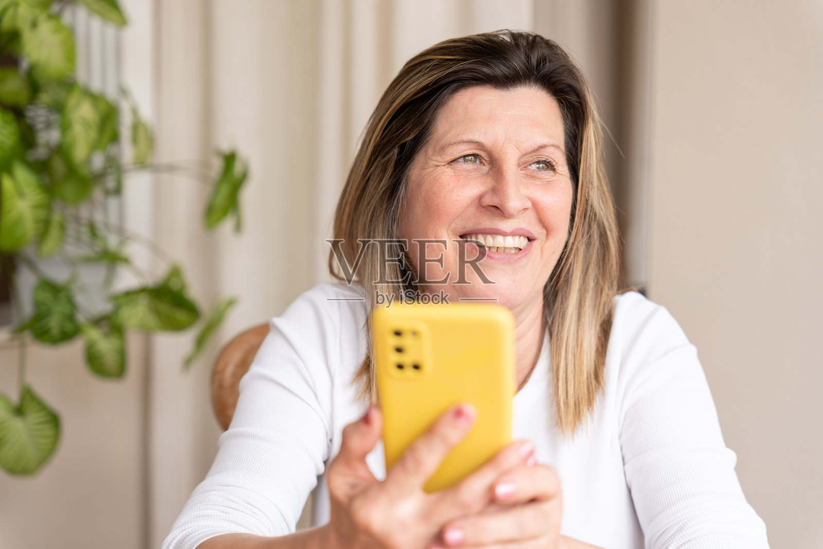 快乐的中年女性，使用黄色智能手机时看向别处，与他人交流照片摄影图片