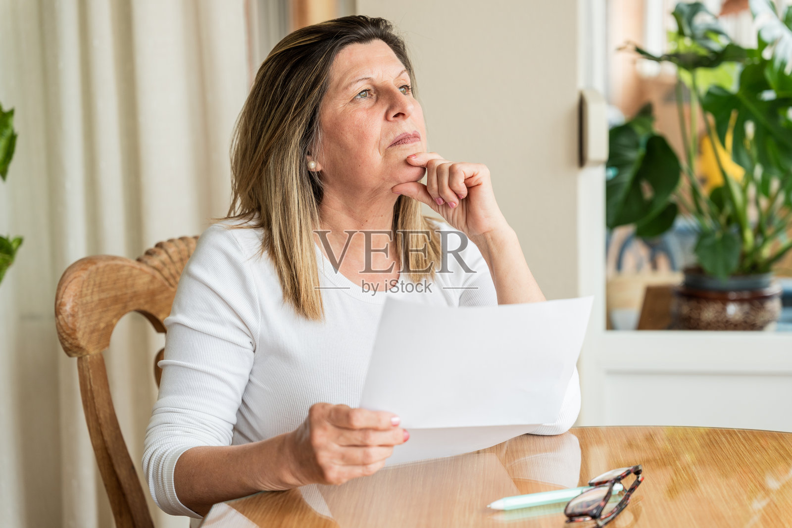 一位成熟的成年女性，手持文件，在木桌旁显得忧虑，似乎在思考财务问题或账单。照片摄影图片