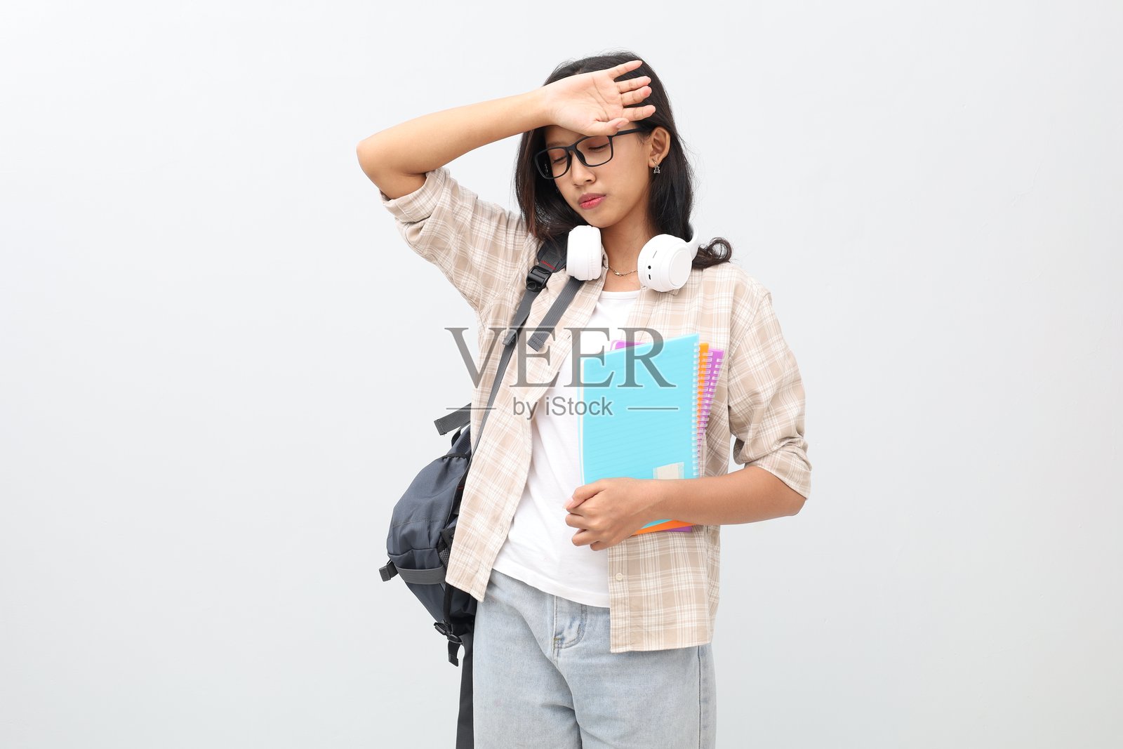 一个疲惫的年轻亚裔女学生，穿着休闲服，戴着眼镜，背着背包，脖子上挂着耳机，手里拿着一本书，孤立在白色背景上，她揉着额头。照片摄影图片