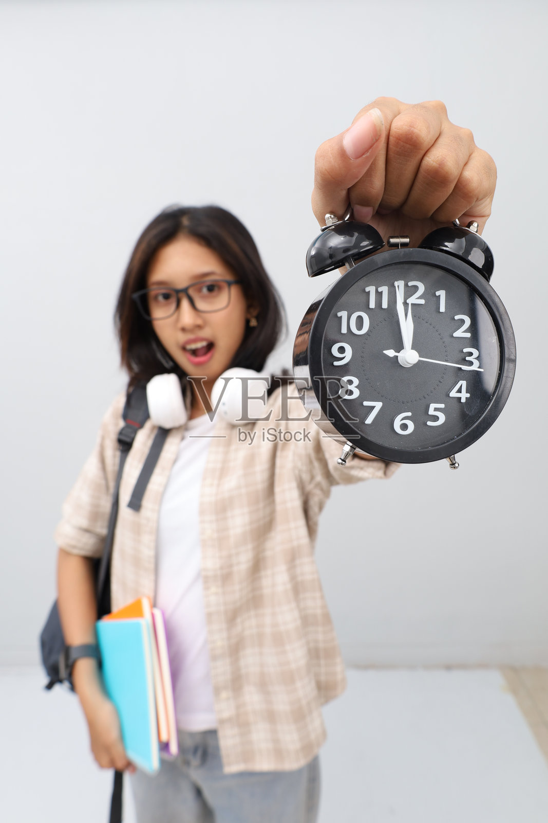 戴眼镜的亚洲女学生，身穿休闲服，背着背包，脖子上挂着耳机，一手拿着书，一手展示着闹钟，脸上露出惊讶的表情看着镜头。照片摄影图片