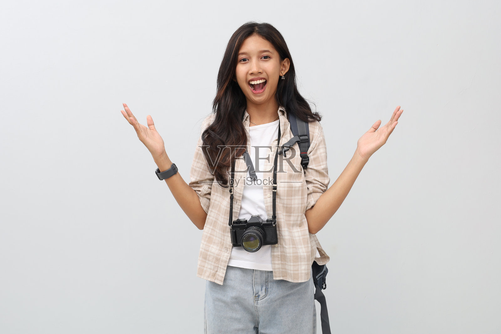 一位年轻亚洲女性穿着休闲服，背着背包，脖子上挂着相机，双手举起，喜悦地尖叫，看起来惊讶于某事，度假概念。照片摄影图片