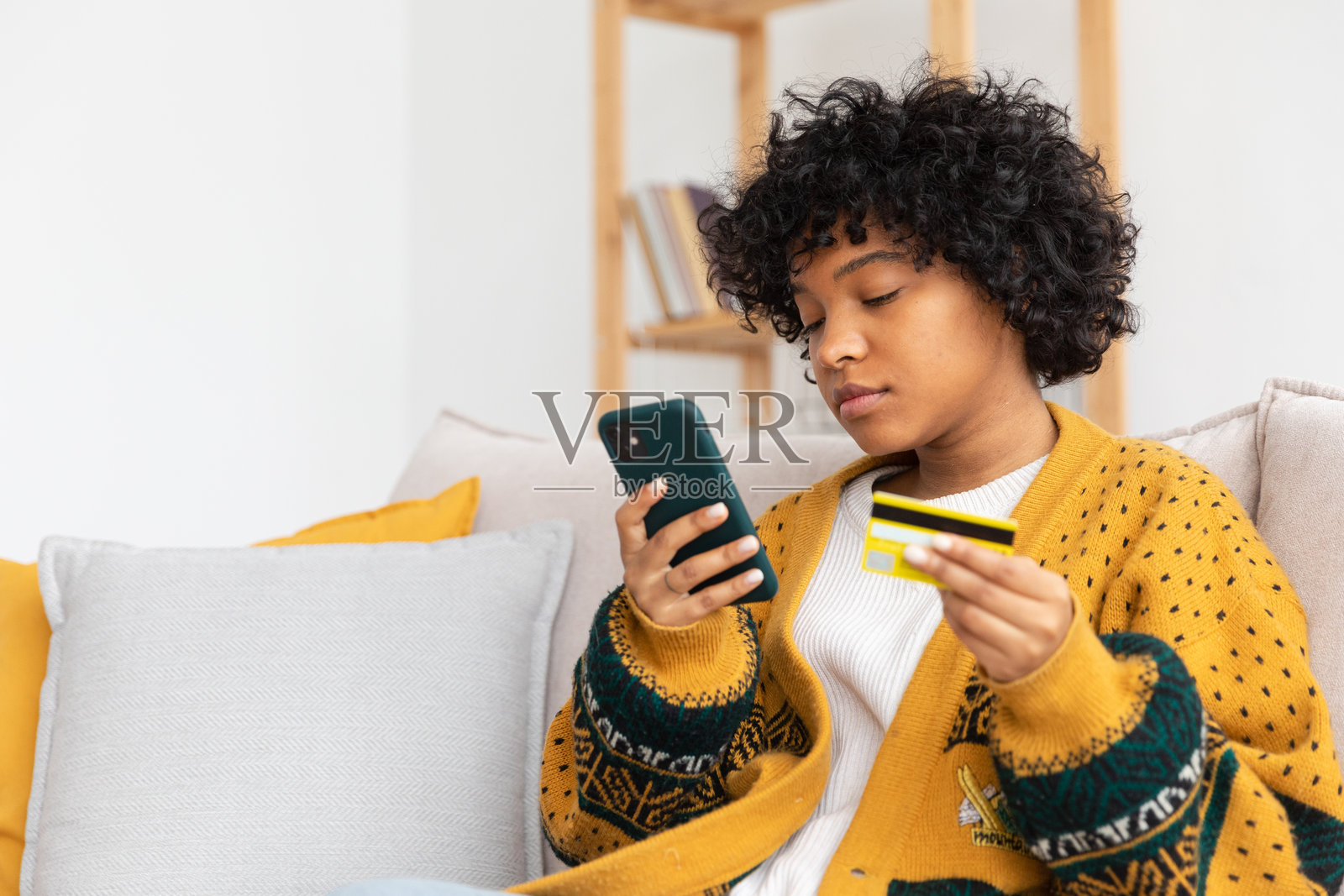 非洲裔美国女性在家中手持智能手机，使用金色信用卡在线购物并输入支付信息。电子商务、网上购物、送货服务。照片摄影图片