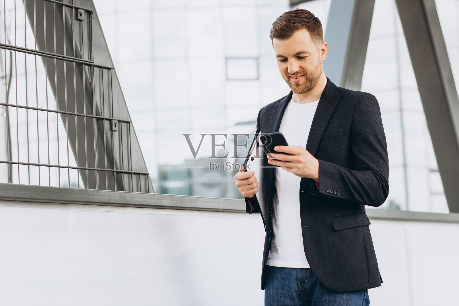 年轻男子站在现代办公楼外，微笑着使用智能手机，享受科技带来的便利照片摄影图片