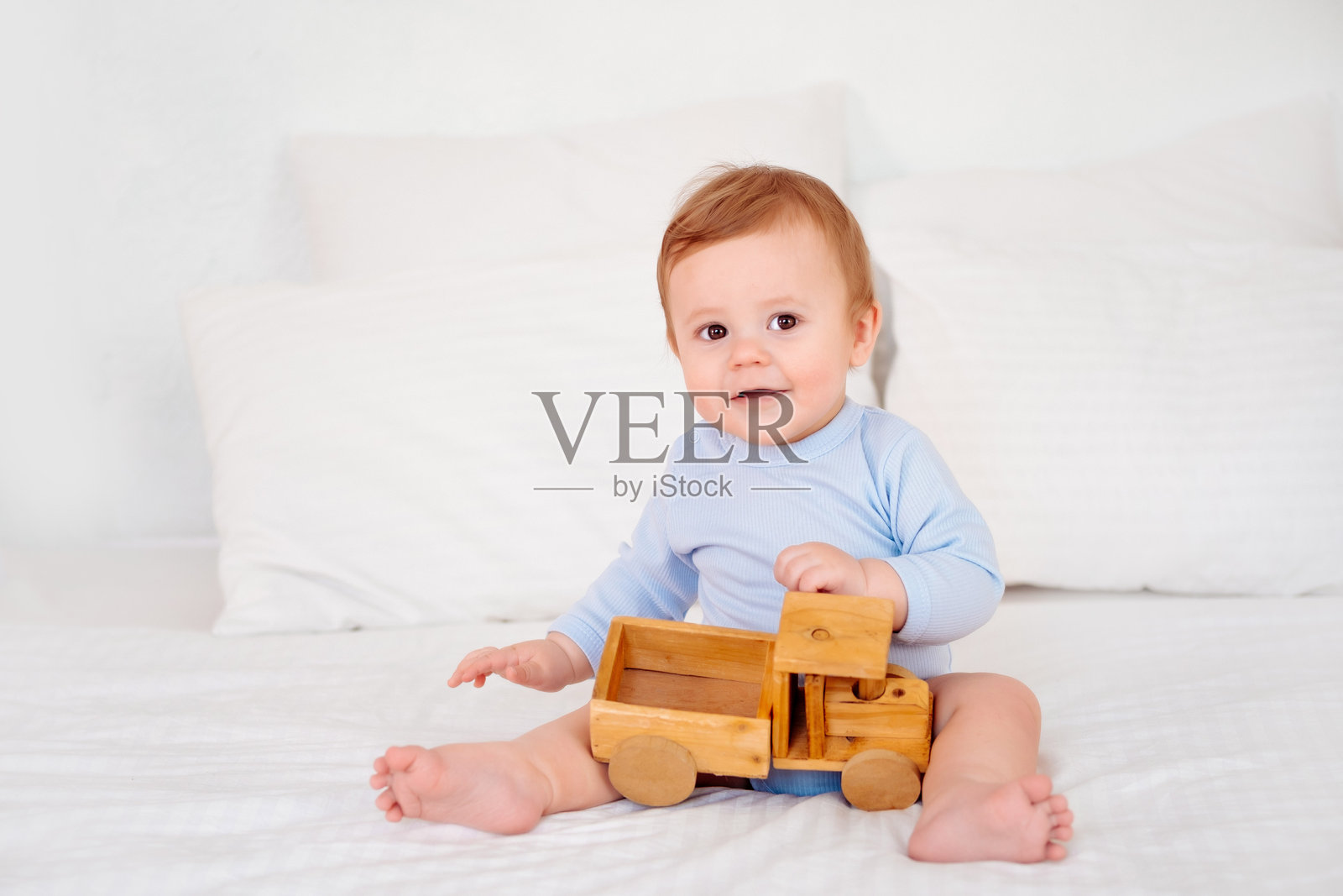 一个孩子在床上玩木制玩具车，一个穿着蓝色连体衣的快乐小童在白色的床上玩耍照片摄影图片