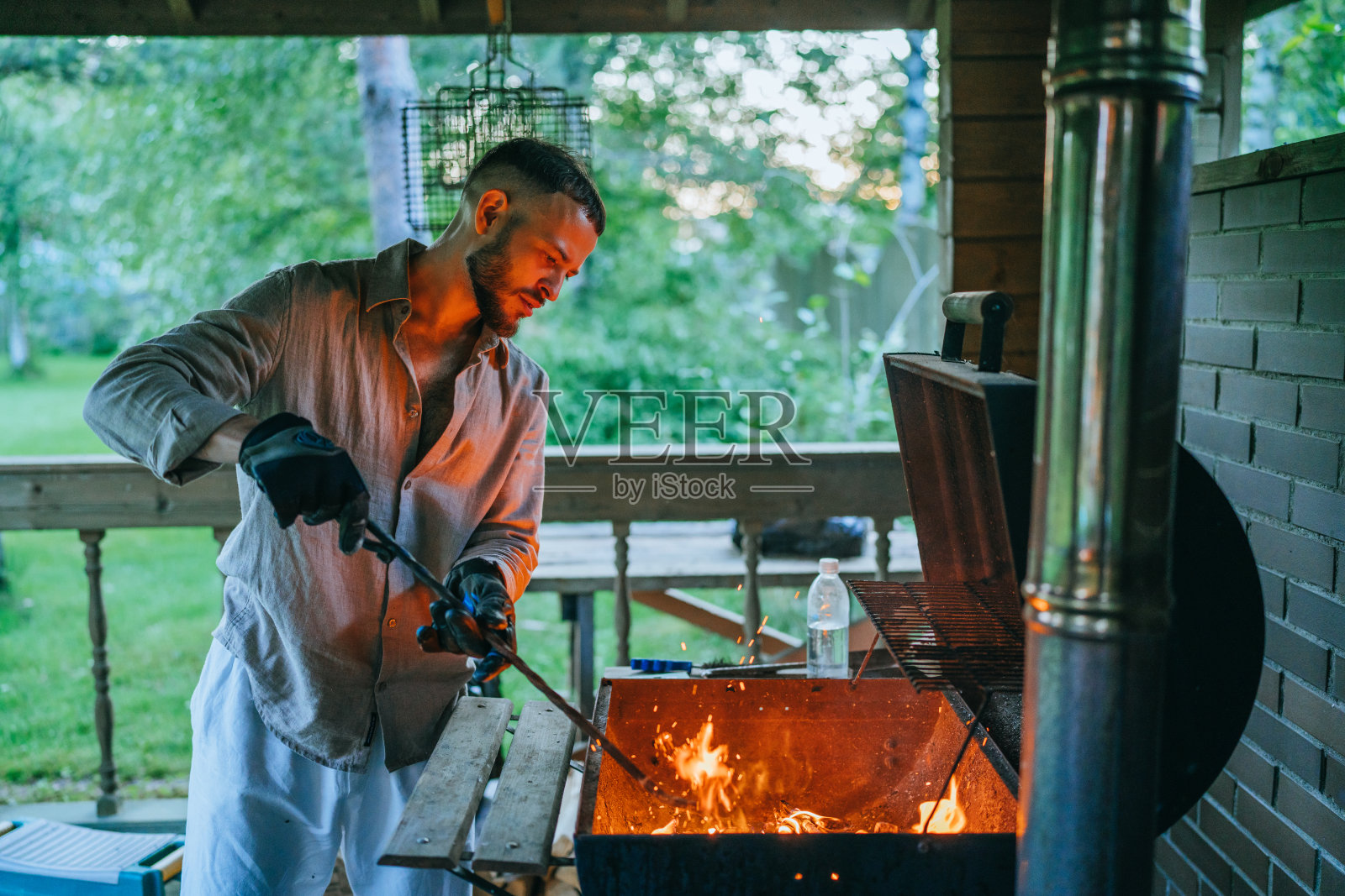 夕阳暖光下，男人专注地在舒适的木制凉亭中烧烤，周围绿意盎然，烤架下有闪烁的余烬。照片摄影图片