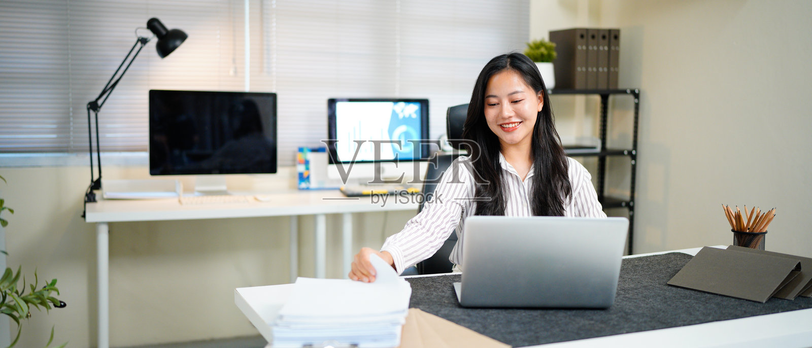 一位商务女性正在繁忙的办公桌上使用笔记本电脑工作，旁边堆放着需要搜索和查阅的纸质文件和文件夹。照片摄影图片