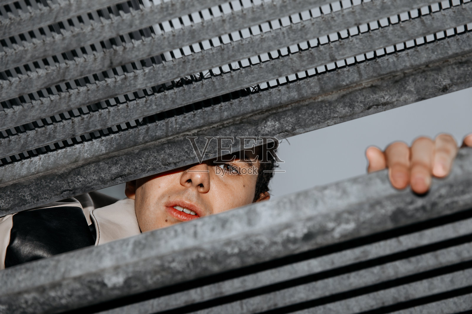 一名年轻的高加索男性带着好奇的表情，透过金属格栅向外窥视照片摄影图片