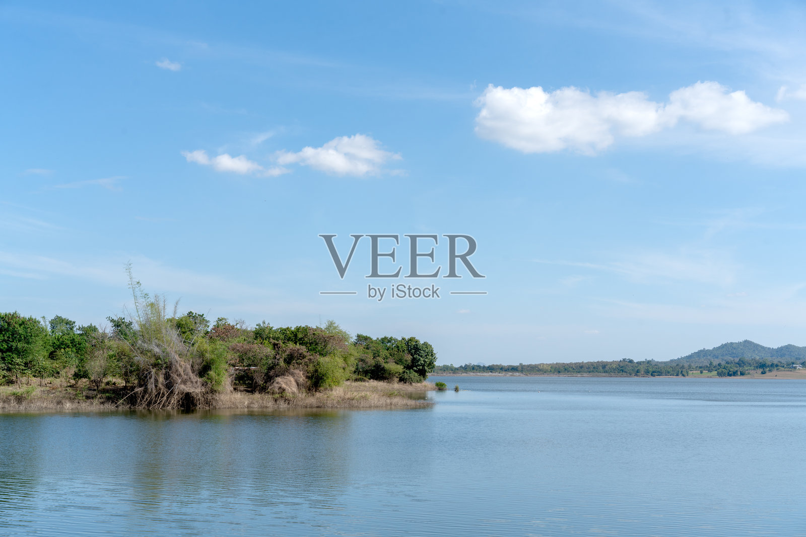 晴朗明亮的天空下，平静的蓝色湖泊，点缀着蓬松的白云。岸边绿树成荫，植被茂密，远方地平线上有连绵的山峦。宁静的自然背景。照片摄影图片