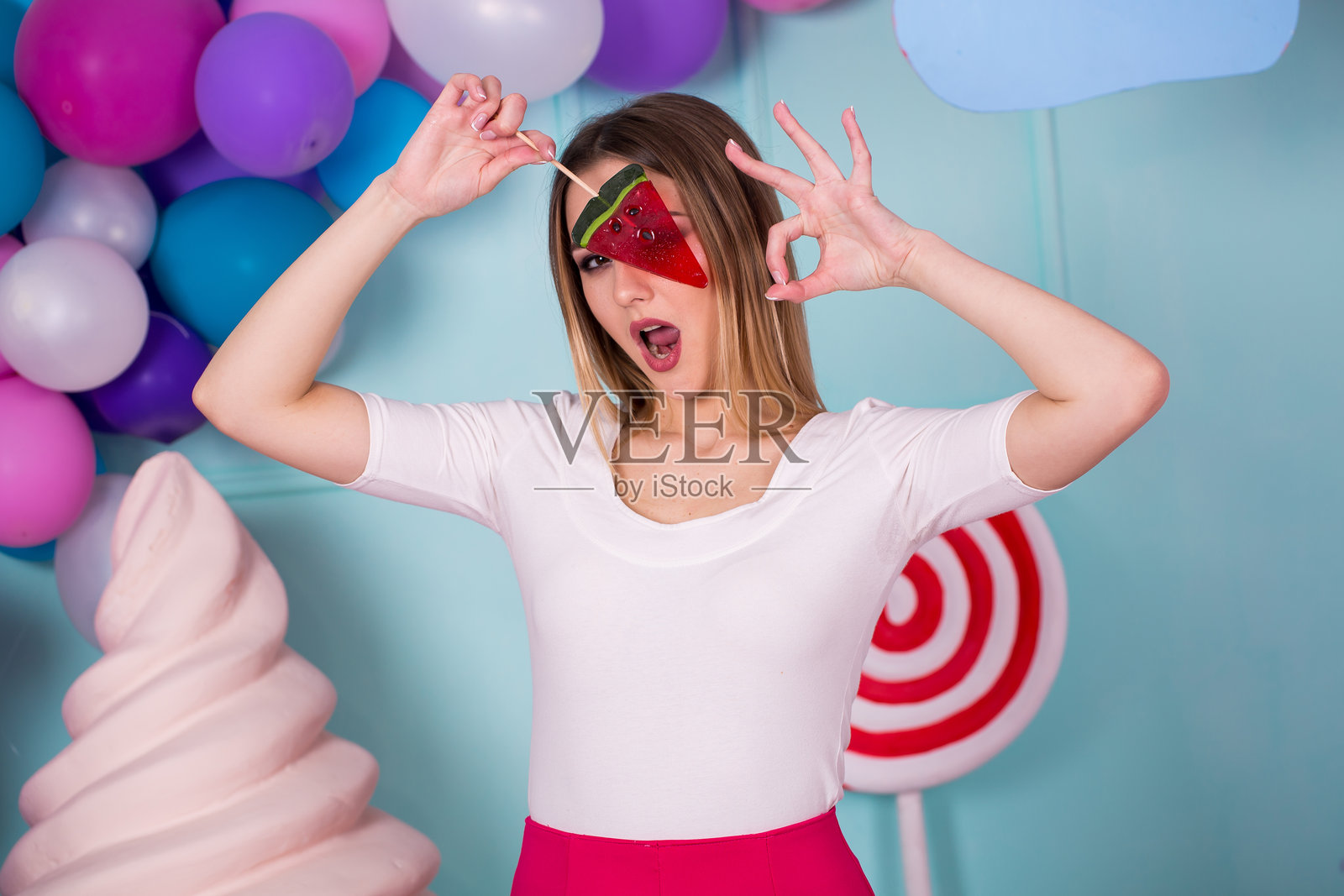 粉色连衣裙的甜食爱好者女士肖像，手持糖果，在装饰着巨型冰淇淋的背景前摆姿势。棒棒糖西瓜。照片摄影图片