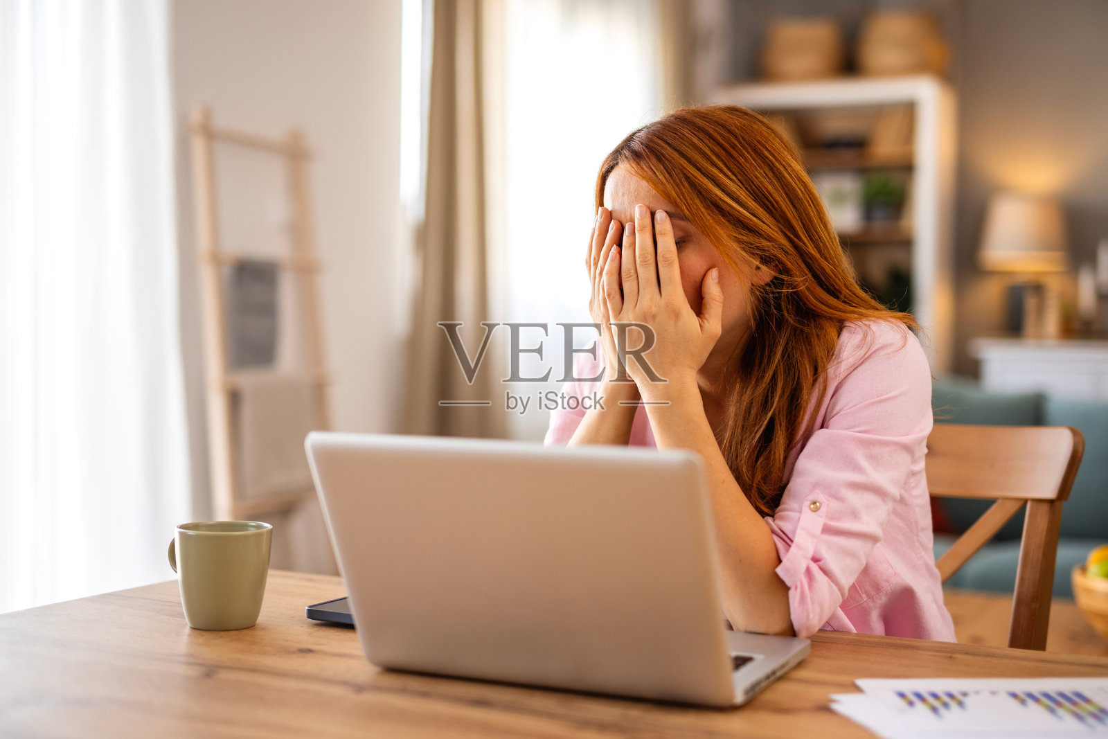 一位疲惫不堪的年轻女子在家中用手捂住脸，同时使用笔记本电脑工作。一位精疲力尽的女自由职业者因过度劳累和压力巨大的截止日期而感到职业倦怠或头痛。照片摄影图片