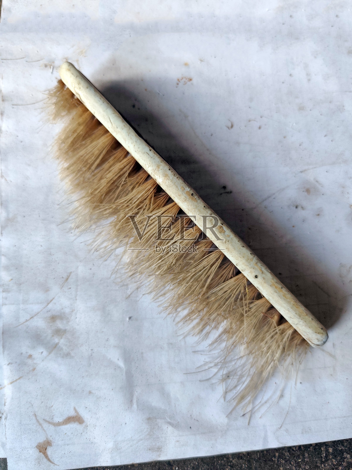 一支带有浅色刷毛、磨损手柄的旧刷子，放在肮脏的白色表面上。这把用过的清洁刷带有明显的污渍、划痕和粗糙的纹理。照片摄影图片