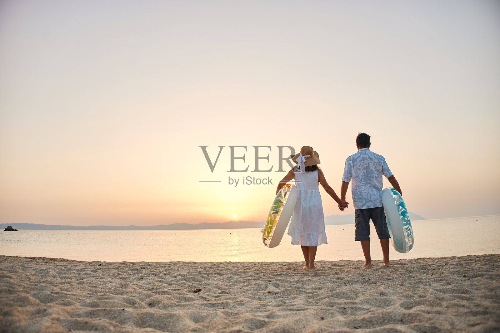 一对情侣牵手漫步在日落海滩，手中提着游泳圈照片摄影图片