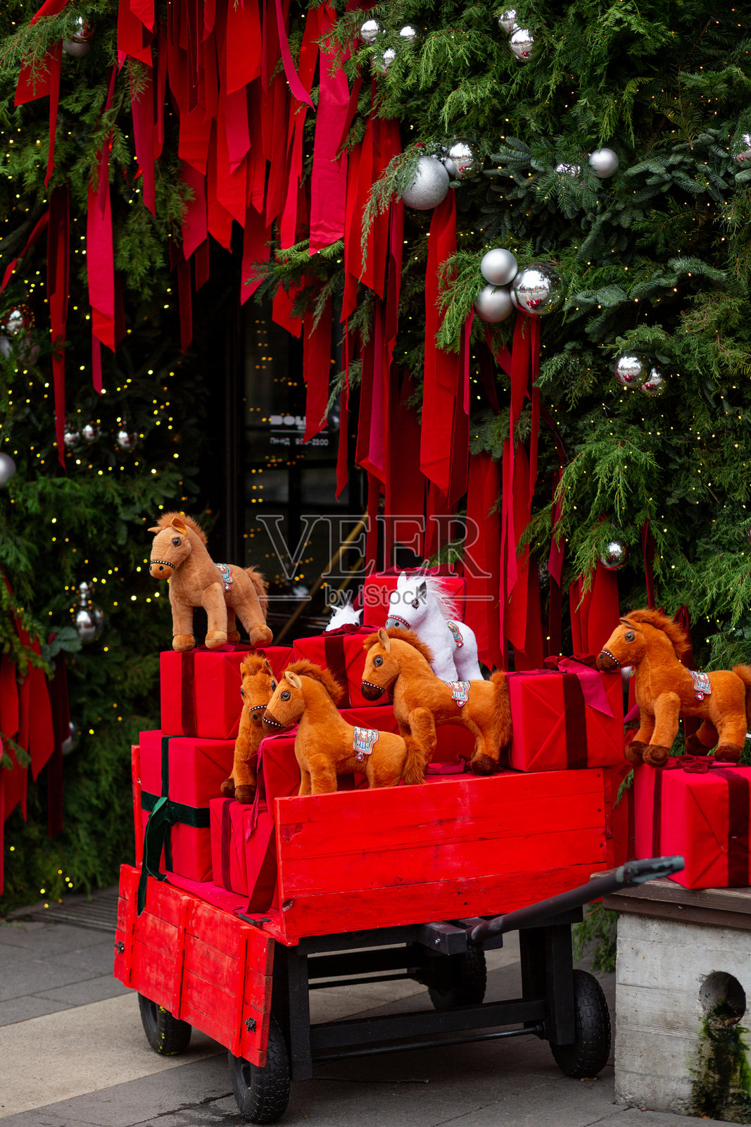 毛绒棕白马匹，装在红色马车里，马车上载有礼品盒，背景是装饰有银色球和花环的松针。照片摄影图片
