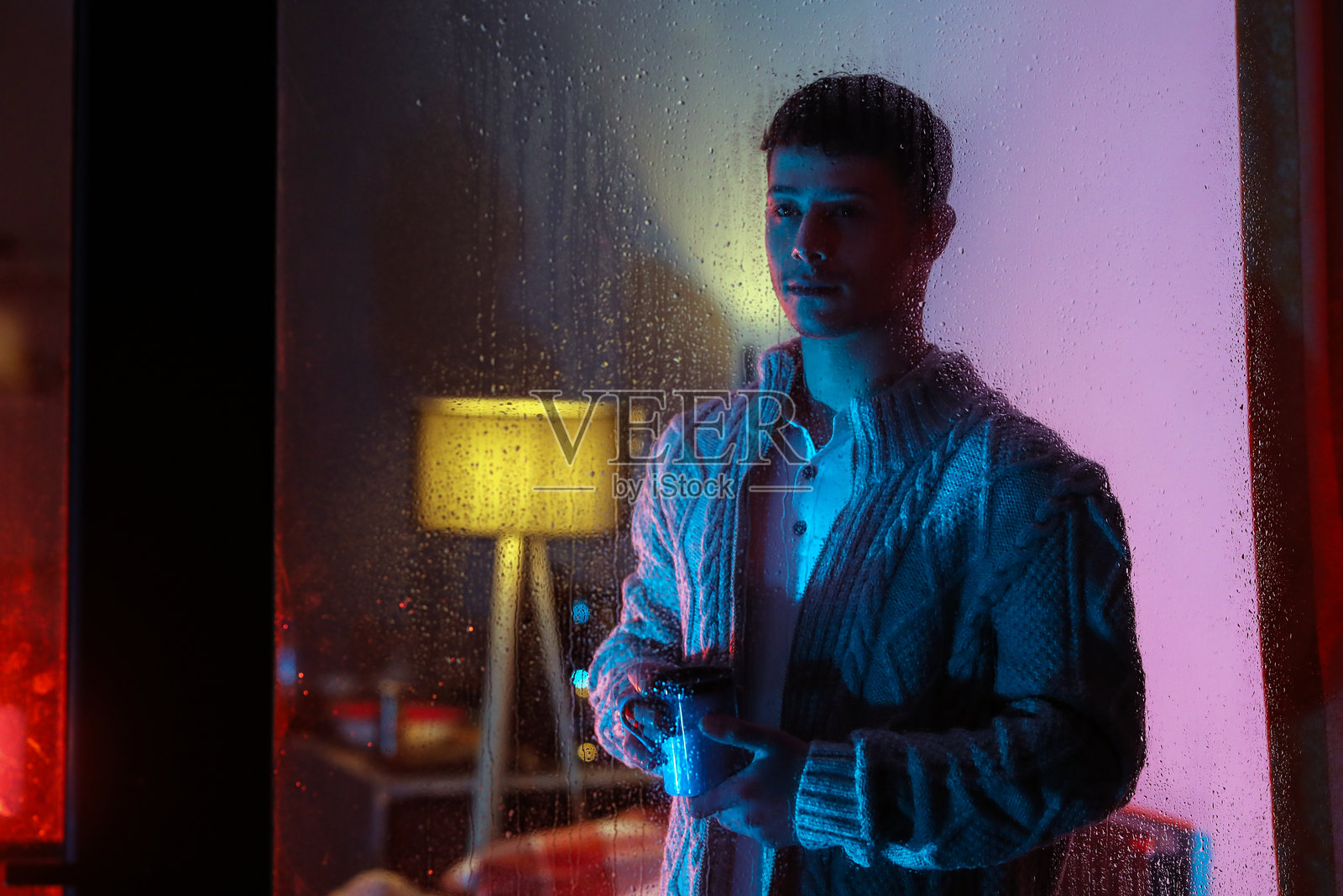 年轻男子在家中，站在雨天的窗边，手持一杯热饮，背景有电影感的彩色灯光照片摄影图片