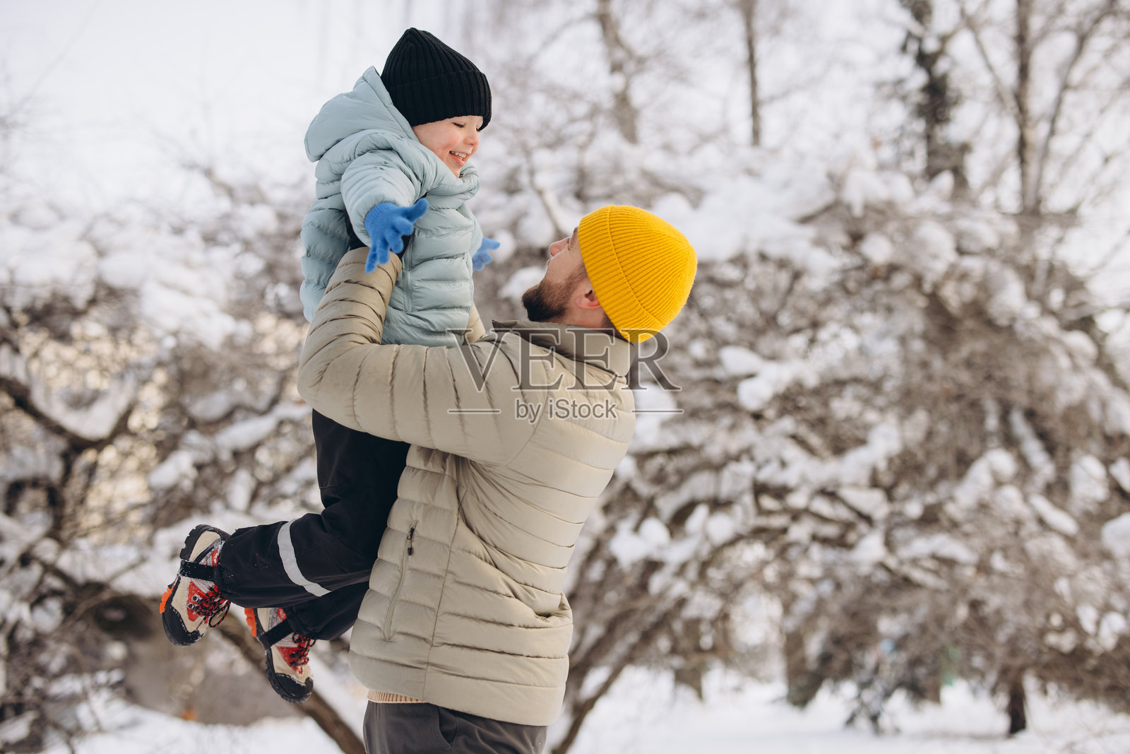 父亲将年幼的儿子高高举起，两人都在雪地冬景中开心地笑着互动。照片摄影图片