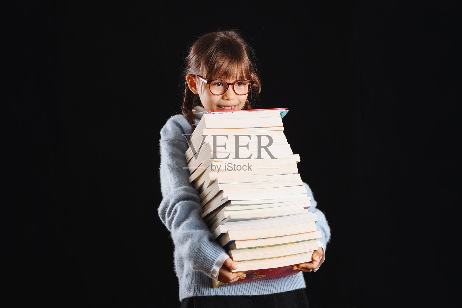 小女孩在身前抱着一大摞书，在影棚的黑色背景前拍摄照片摄影图片