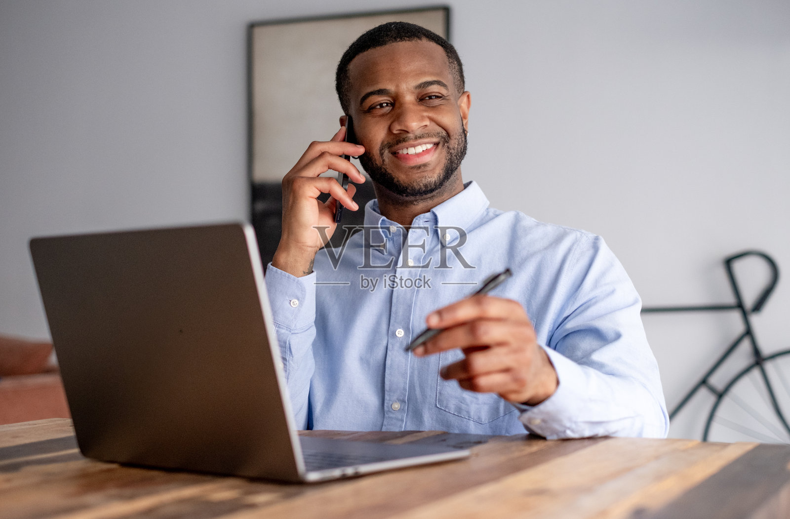 一位职业非洲裔美国男性公司员工的肖像，年轻的微笑男商人、创业者或工人在木桌上用笔记本电脑打电话。照片摄影图片