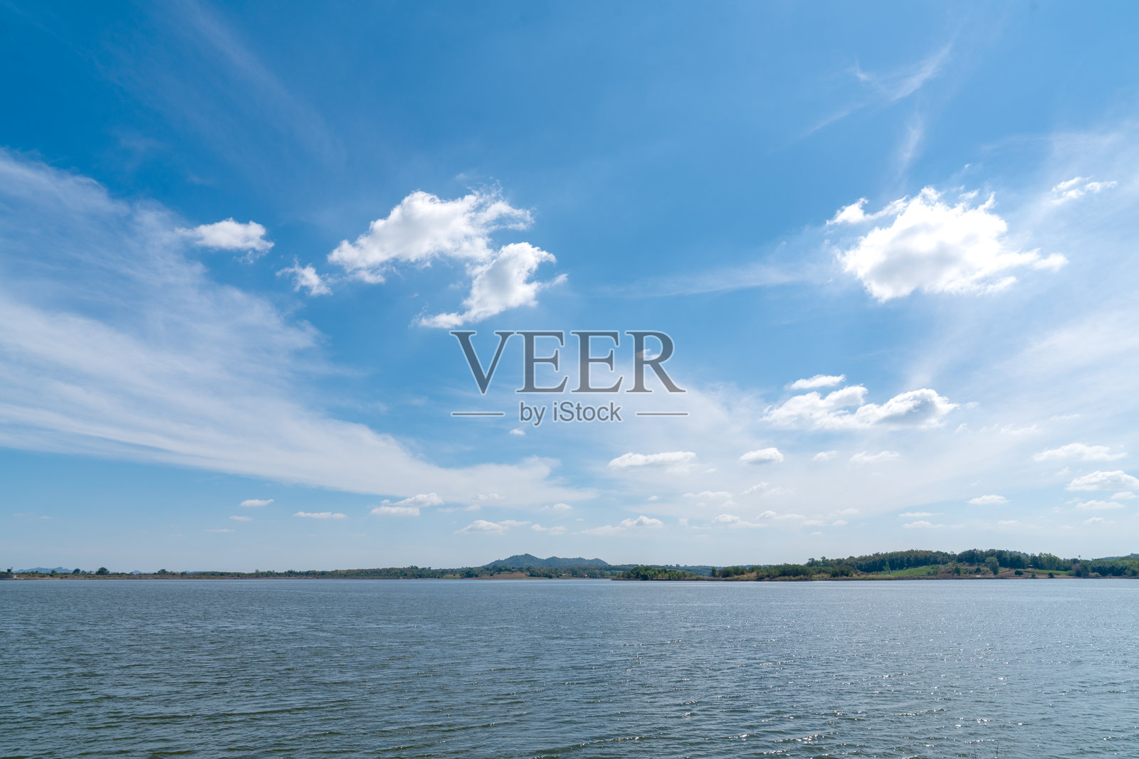 阳光明媚的晴空下，白色的卷云飘浮，一汪平静的蓝色湖水构成宽广的风景。远处绿色的山丘和树林环绕着水库，景色宜人，是宁静的户外自然背景。照片摄影图片