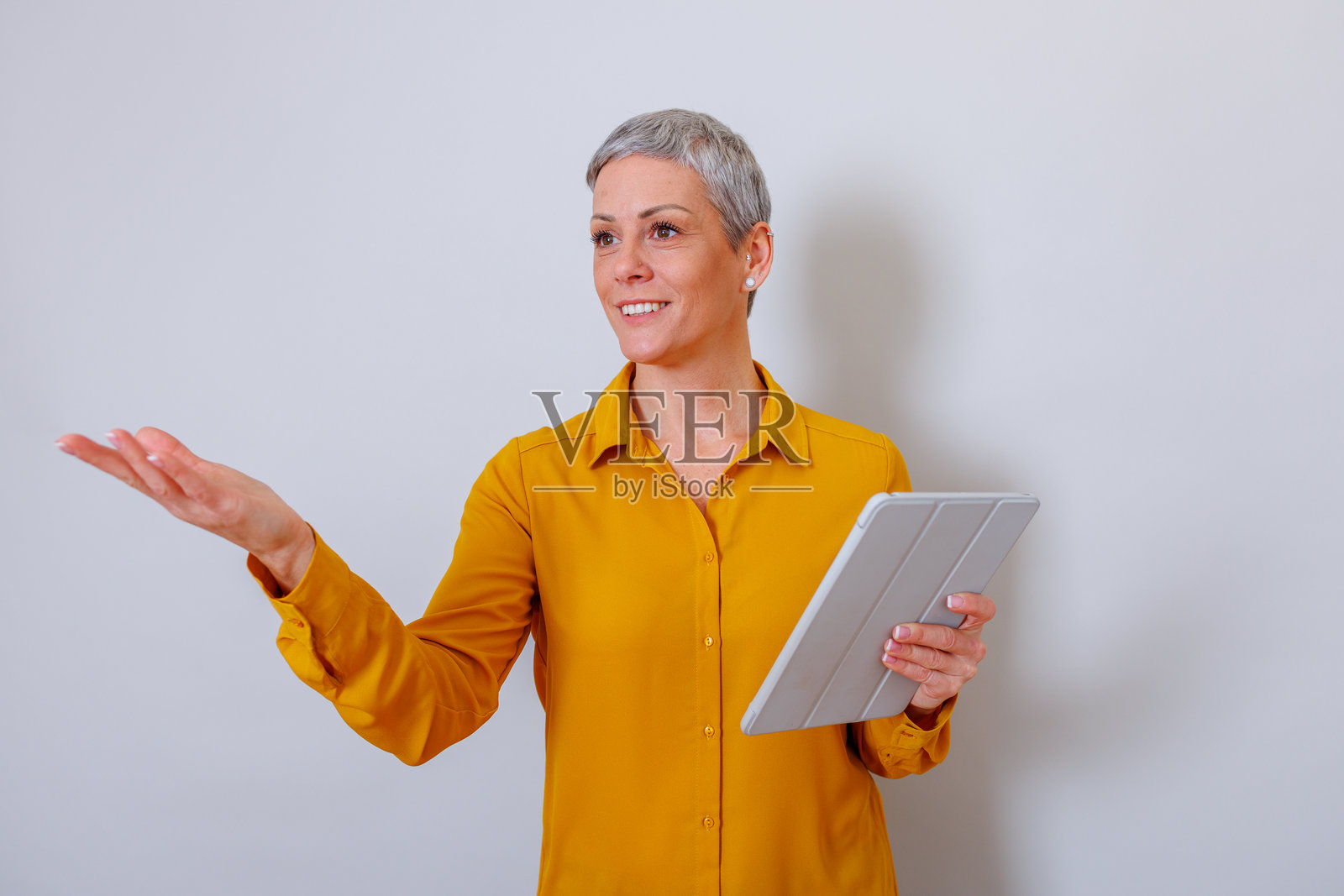 一位中年女性，留着灰色短发，面带微笑，手持平板电脑，向前伸出手指进行展示。照片摄影图片