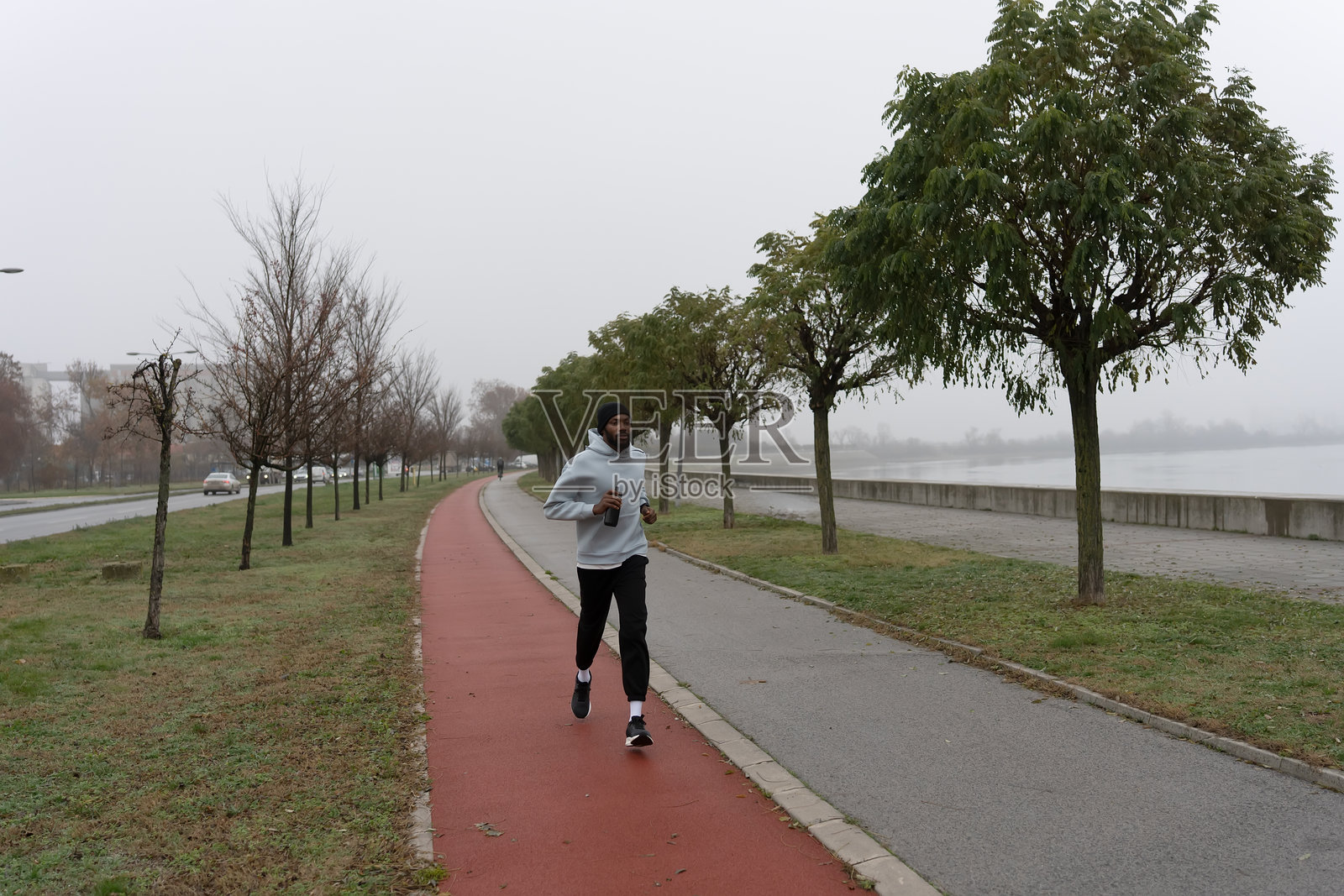 年轻的运动男子在河畔林荫小道慢跑，身穿连帽衫和绒线帽。城市跑步锻炼、耐力训练、健康积极的都市生活方式照片摄影图片