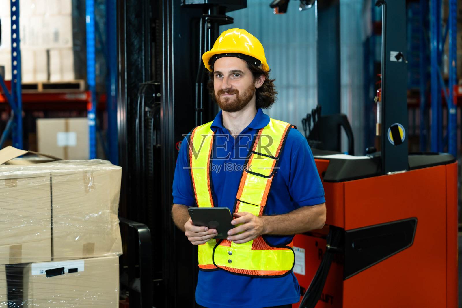 仓库工作人员在物流部门的盘点作业中，使用数字平板电脑，站在堆叠的包裹和叉车旁边，位于一个井然有序的仓储中心内，身处物流仓库之中。照片摄影图片
