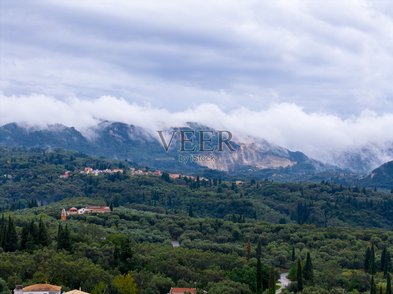 科孚岛风景如画的希腊景色，橄榄树林山谷中坐落着一个村庄，远处的山峦笼罩在薄雾中，云层密布的天空下可见一个大型采石场。照片摄影图片