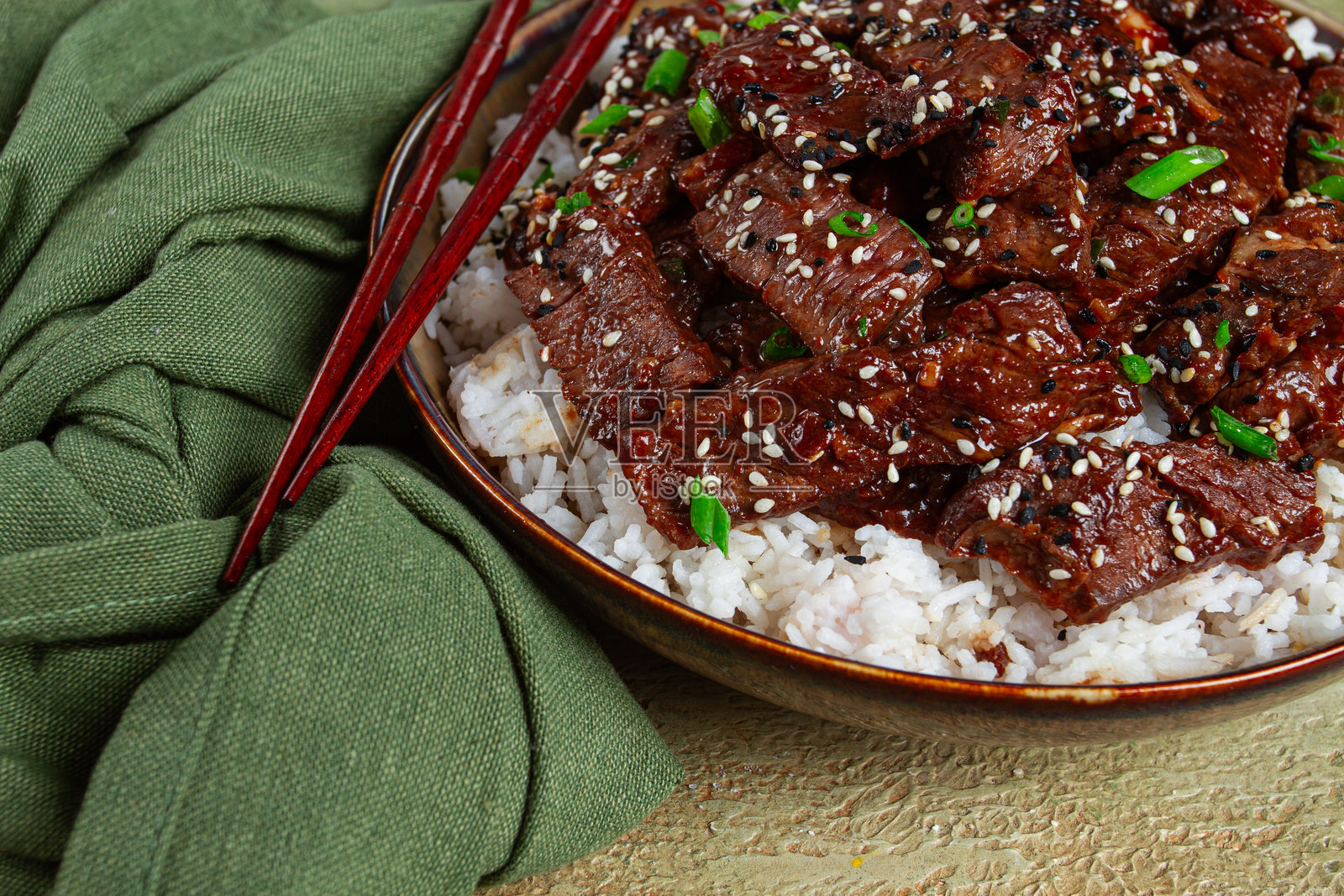 脆皮牛肉，配以葱花和芝麻，盛放在米饭垫上，装在碗里，配有筷子，放在纹理表面上，无人像照片摄影图片