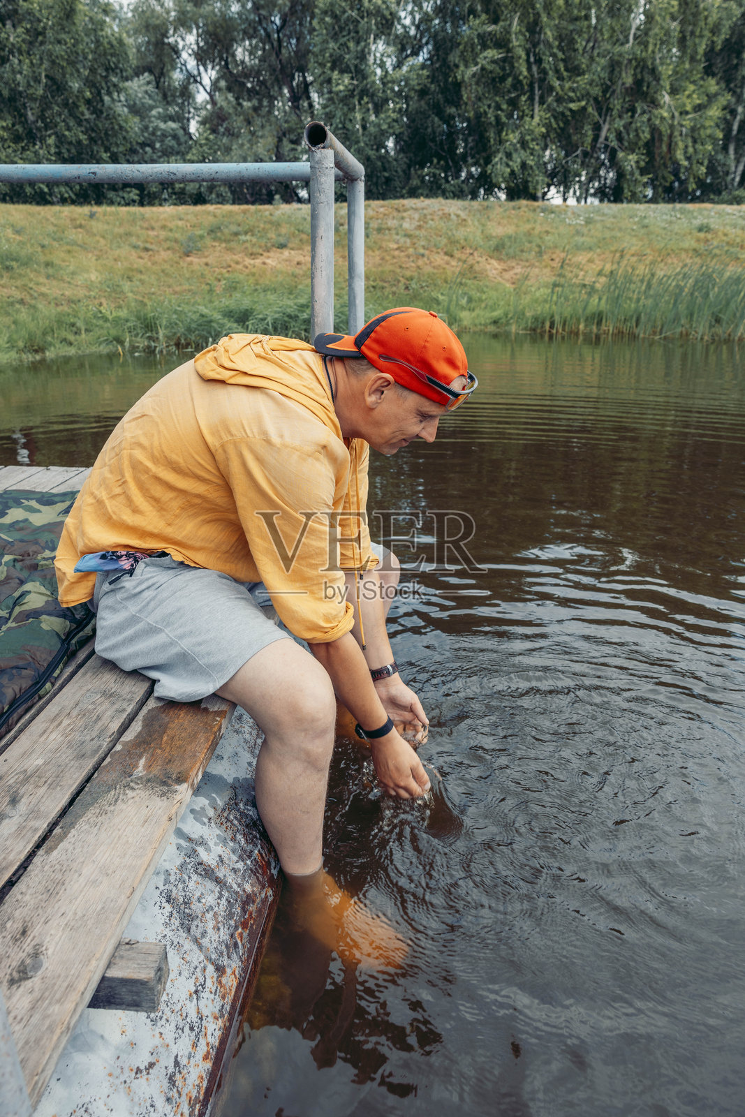 一个年轻的高加索男子，留着短发，戴着红色帽子，坐在码头上，双脚浸在被绿植环绕的平静湖水中。这是一个晴朗的日子。照片摄影图片