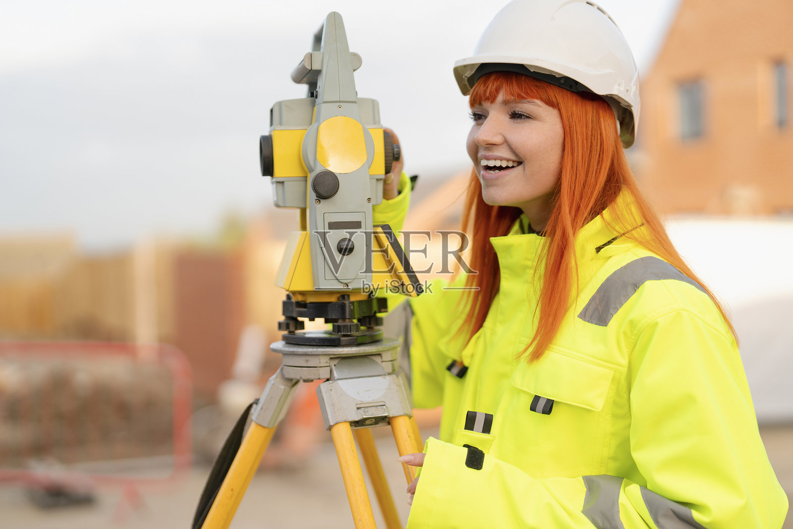 身着亮黄色安全装备、笑容甜美的女性测量专家在建筑工地使用全站仪，她留着一头红发照片摄影图片