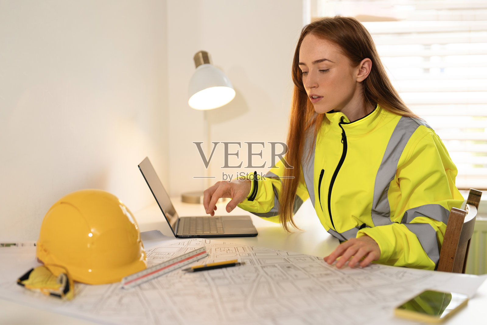 一位迷人的长发女性建筑专业人士在明亮的工作空间里，一边使用笔记本电脑一边分析图纸照片摄影图片