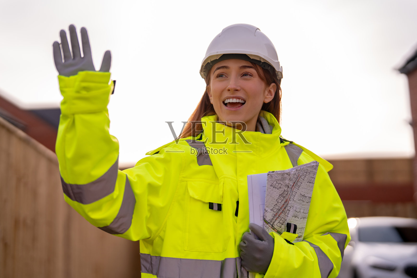 年轻的女建筑工，留着长发，在建筑工地上微笑着挥手，手中拿着建筑图纸。照片摄影图片