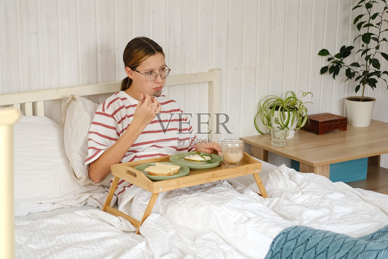 戴眼镜的少女在床上吃早餐，喝着茶，在家享受早晨时光。少女在床上的托盘里吃东西。穿着睡衣的少女在卧室里放松地喝着咖啡。照片摄影图片