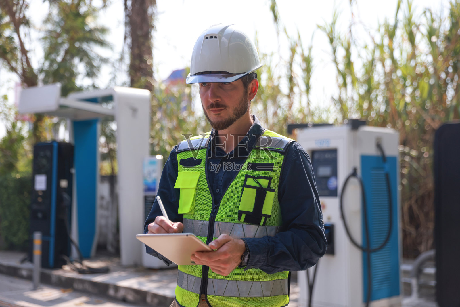 一名戴着安全帽和高能见度背心的工业工程师或电气工程师在户外的电动汽车充电站使用平板电脑，展示电动汽车技术和服务行业。照片摄影图片