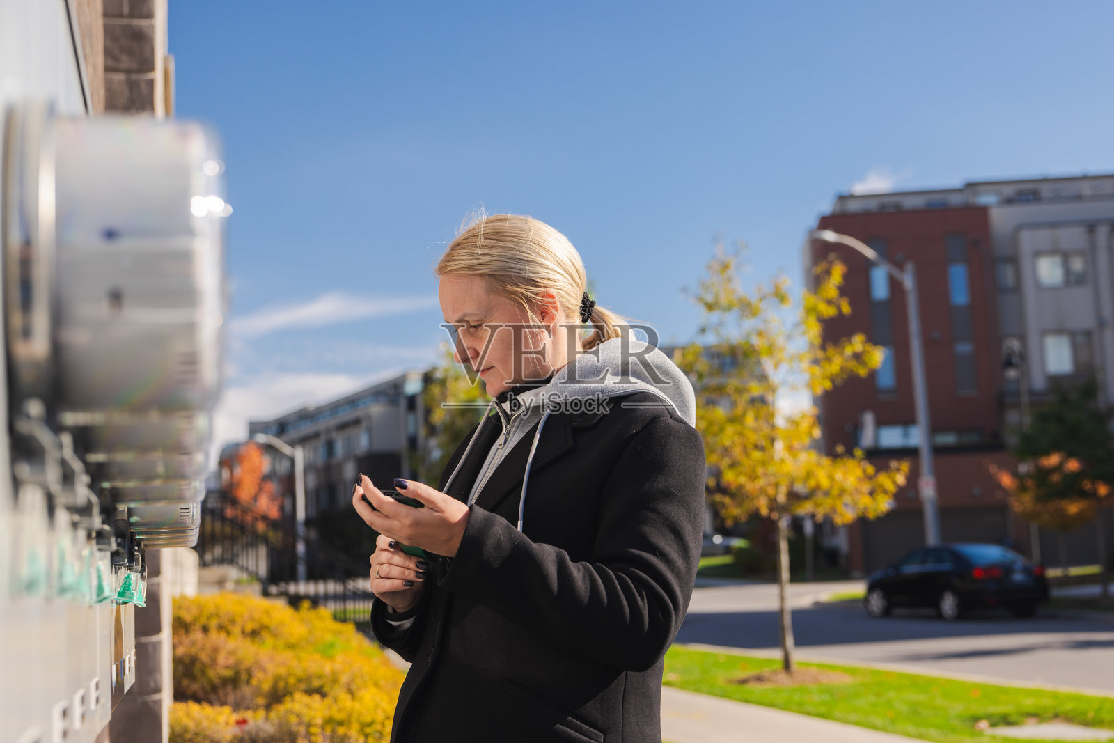 秋日晴朗，一位中年女性在住宅区读取户外电表，并用智能手机记录用量数据。照片摄影图片